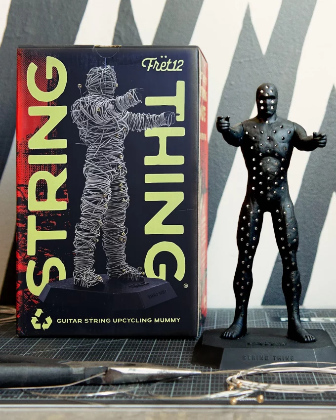 A black plastic mummy figurine called the String Thing designed for upcycling old guitar strings, featuring a perforated body for weaving and a FRET12 logo base.