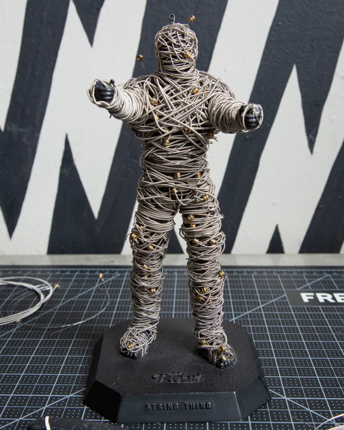 A black plastic mummy figurine called the String Thing designed for upcycling old guitar strings, featuring a perforated body for weaving and a FRET12 logo base.