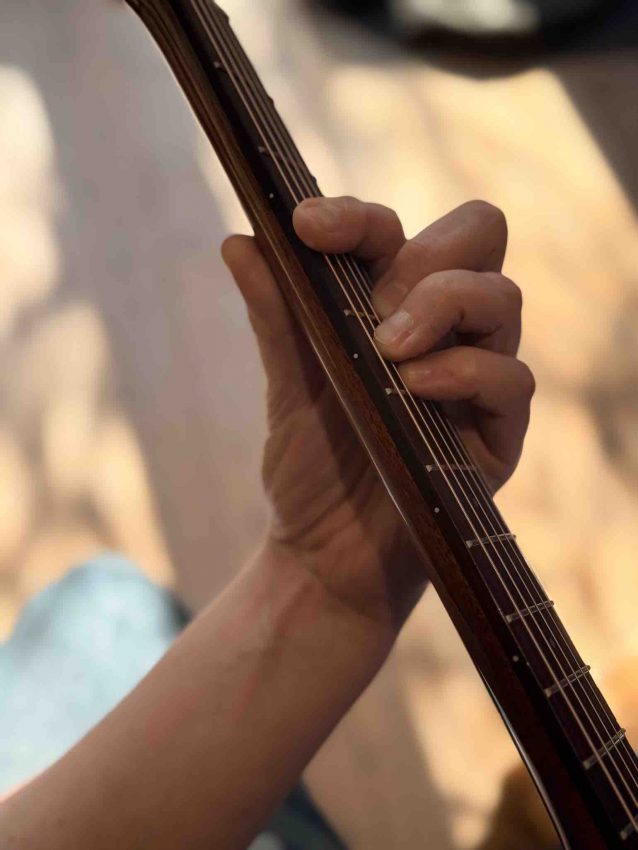 Close-up of a guitarist's fretting hand showing the index finger rolled slightly on its side to use the bony edge for a barre chord on an acoustic guitar.