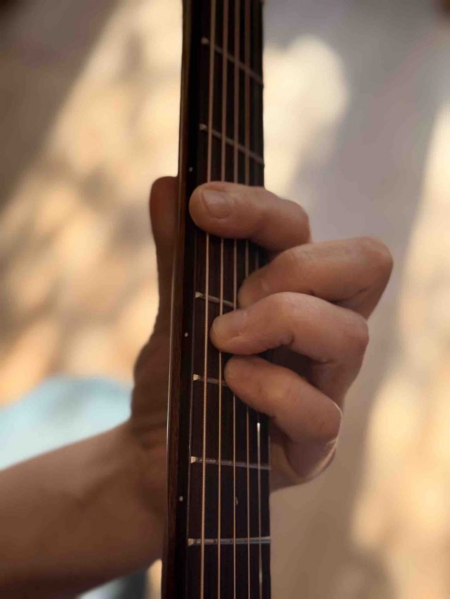 Close-up of a guitarist's fretting hand showing the index finger rolled slightly on its side to use the bony edge for a barre chord on an acoustic guitar.