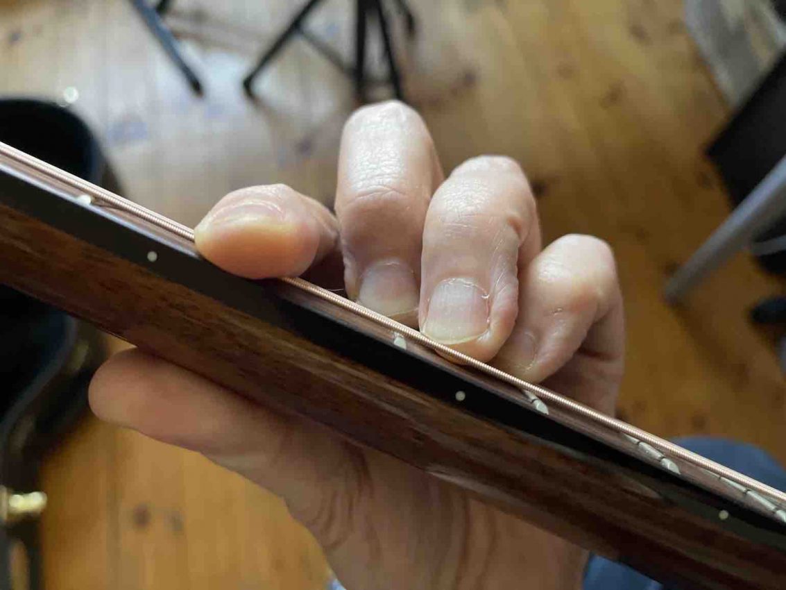 Close-up of a guitarist's fretting hand showing the index finger rolled slightly on its side to use the bony edge for a barre chord on an acoustic guitar.