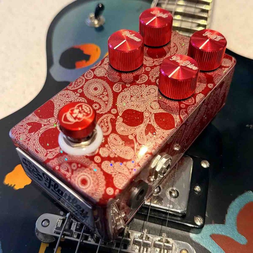 A high-angle shot of the Flattley JK Fuzz pedal, featuring its unique hand-processed finish, custom red anodized aluminum control knobs, and a red halo light ring on a premium boutique guitar effects unit.